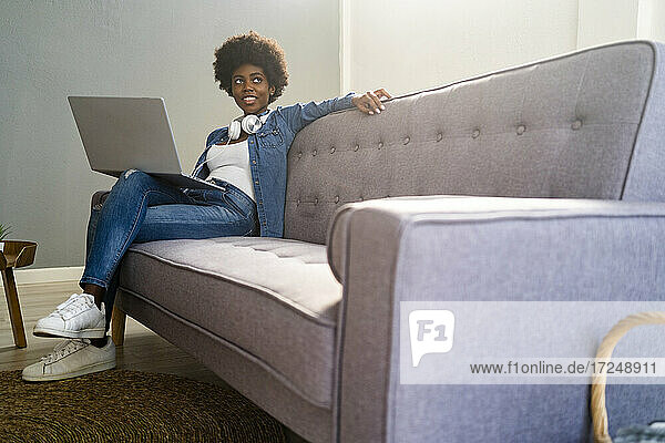 Junge Frau  die wegschaut  während sie mit ihrem Laptop auf dem Sofa zu Hause sitzt