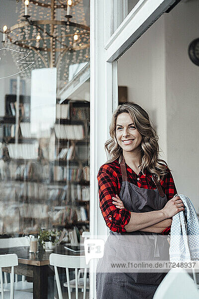 Confident female owner with arms crossed leaning on door at cafe