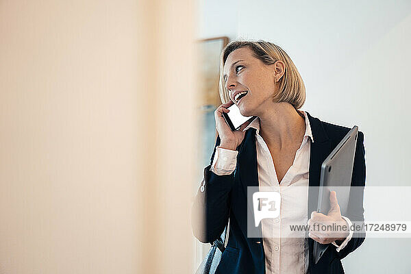 Smiling businesswoman holding laptop while talking on phone at home