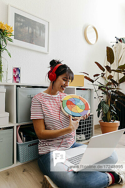 Frau mit Kopfhörern  die bei einem Videogespräch über einen Laptop zu Hause eine Farbmusterplatte zeigt