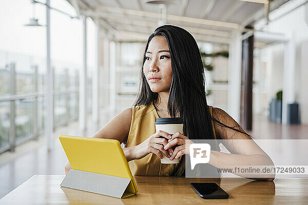 Geschäftsfrau schaut weg  während sie eine Kaffeetasse in einer modernen Büro-Cafeteria hält