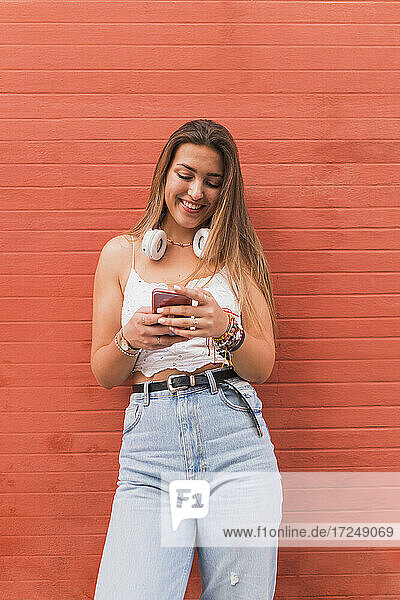 Lächelnde Frau  die ein Mobiltelefon benutzt und vor einer orangefarbenen Wand steht
