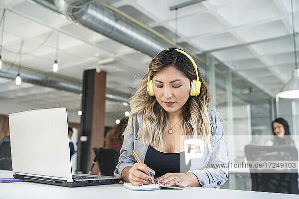 Geschäftsfrau mit Kopfhörern  die am Laptop im Büro in ihr Tagebuch schreibt