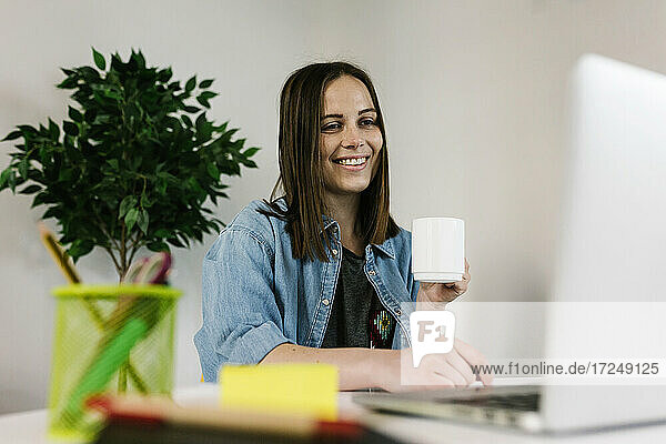 Lächelnde Unternehmerin  die an einem Laptop arbeitet  während sie einen Kaffee am Schreibtisch im Heimbüro trinkt