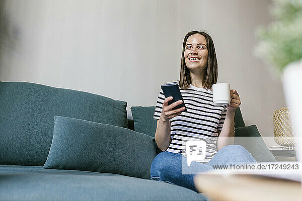 Lächelnde Frau mit Kaffee und Handy in der Hand auf dem Sofa im Ferienhaus