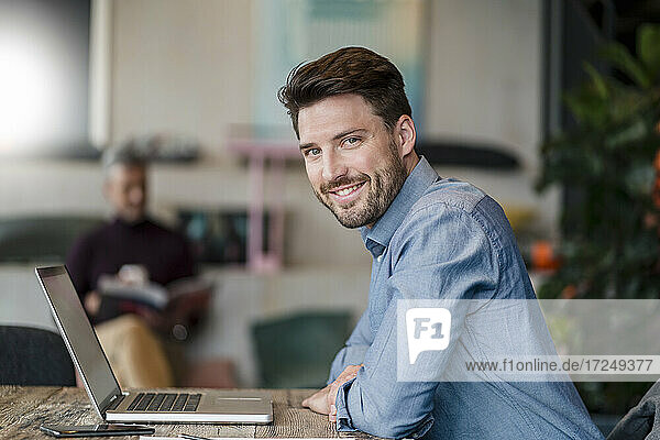Lächelnder männlicher Unternehmer mit Laptop  der sich bei der Arbeit auf einen Tisch stützt