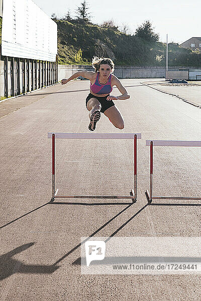 Young female athlete jumping over hurdle during sunny day