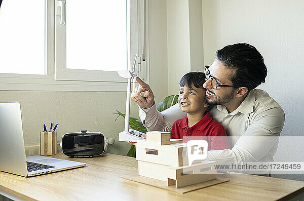 Männlicher Architekt  der auf eine Windkraftanlage zeigt  während er mit seinem Sohn im Büro sitzt