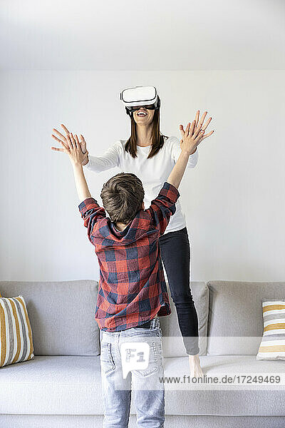 Lächelnde Mutter  die ein virtuelles Headset trägt und mit ihrem Sohn spielt  während sie auf dem Sofa im Wohnzimmer steht