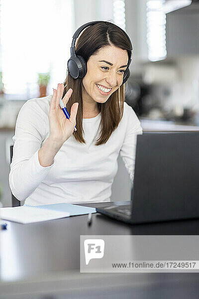 Lächelnde weibliche Fachkraft winkt während einer Videokonferenz über einen Laptop zu Hause