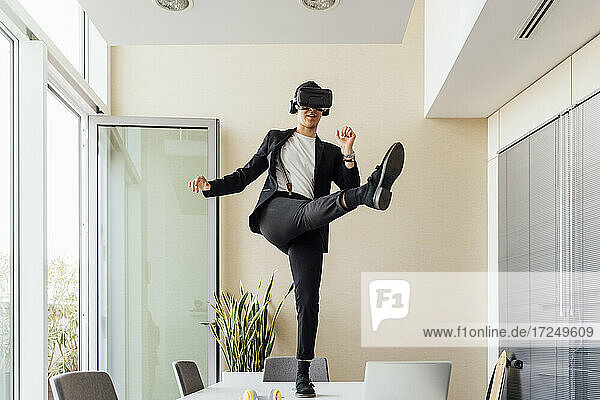 Young female entrepreneur with virtual reality headset kicking while standing on desk in office