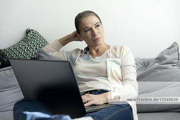 Nachdenkliche Frau sitzt mit Laptop auf dem Sofa zu Hause