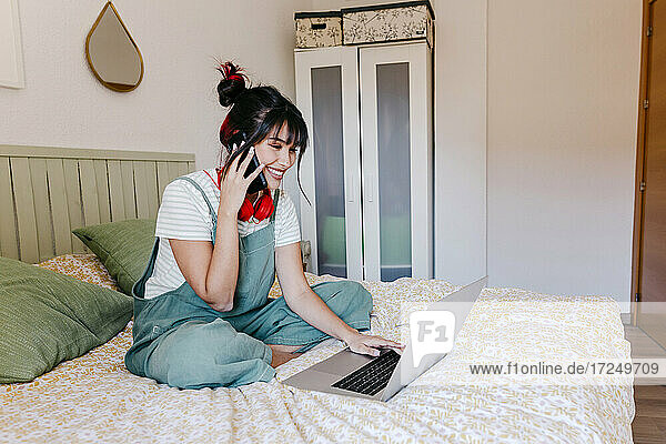 Junge Frau spricht mit dem Handy  während sie einen Laptop im Schlafzimmer benutzt