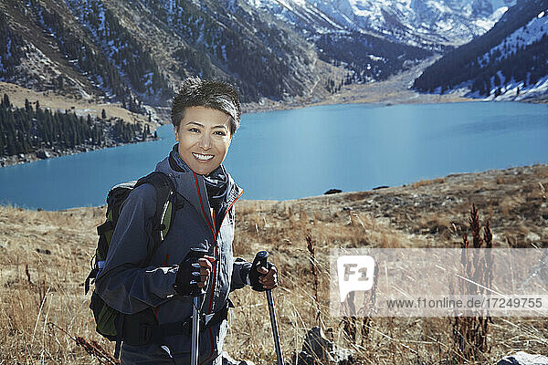 Smiling woman wearing backpack holding hiking poles by lake