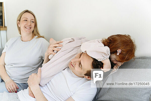 Lächelnde schwangere Frau  die ihre Tochter betrachtet  die mit ihrem Vater auf dem Sofa zu Hause spielt