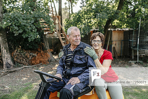 Smiling senior couple sitting on lawn mover in backyard
