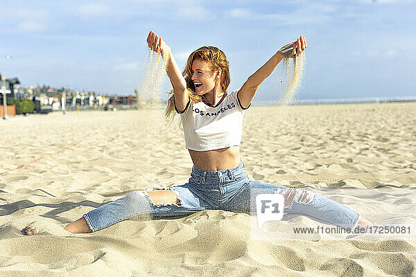 Unbeschwerte junge Frau spielt mit Sand am Strand an einem sonnigen Tag