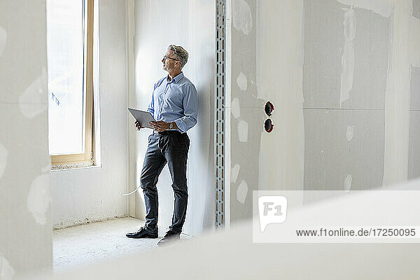 Männlicher Fachmann  der wegschaut und einen Tablet-PC hält  während er an einer Wand auf einer Baustelle steht