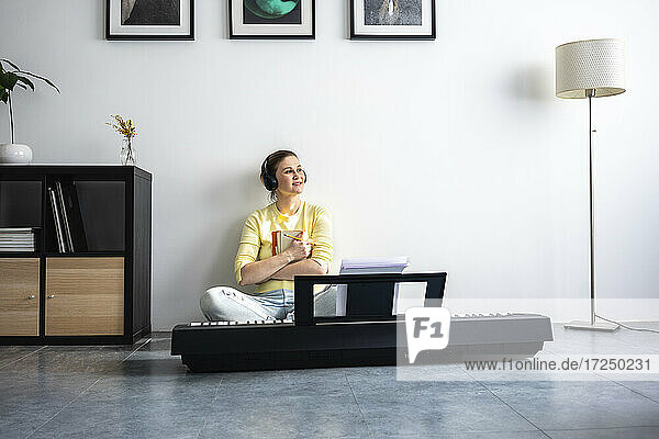 Lächelnde Frau mit In-Ear-Kopfhörern  die ein Buch hält  während sie zu Hause vor einem Klavier sitzt