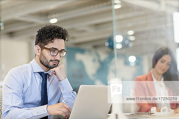 Junger Geschäftsmann mit Brille arbeitet am Laptop mit einer Kollegin im Hintergrund im Büro
