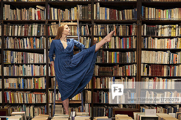Junge Frau in blauem Kleid balanciert auf einem Bein auf einer Leiter in einer Bibliothek