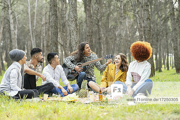 Young male and female friends playing guitar during picnic at forest