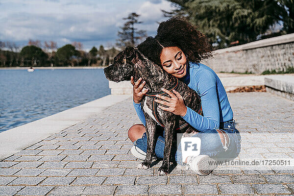 Curly hair woman embracing dog while sitting on footpath