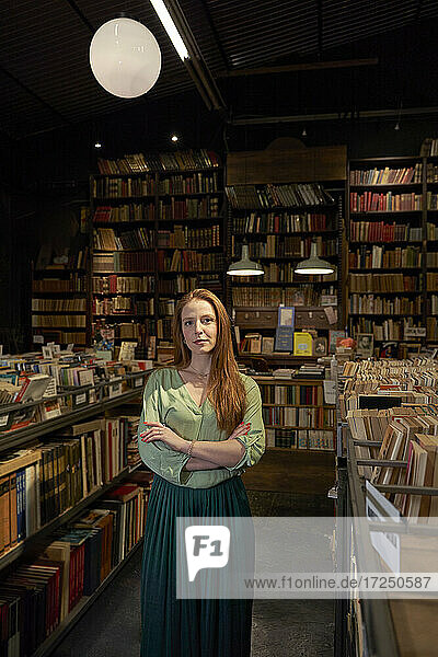 Schöne junge Frau mit verschränkten Armen steht inmitten von Bücherregalen in der Bibliothek