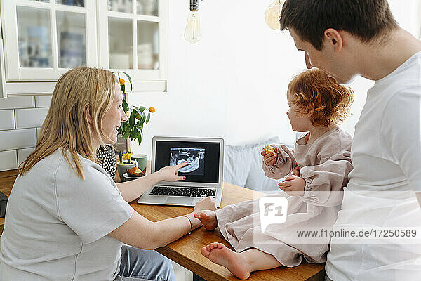 Lächelnde schwangere Frau zeigt ihrer Tochter das Ultraschallbild ihres Vaters auf dem Wohnzimmertisch