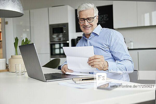 Lächelnder männlicher Unternehmer  der mit einem Laptop im Büro sitzt und ein Dokument liest