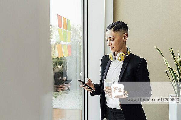Unternehmerin hält Einweg-Kaffeebecher  während sie im Büro ihr Smartphone benutzt
