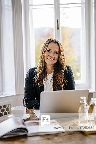 Lächelnde weibliche Fachkraft sitzt mit Laptop am Tisch im Büro