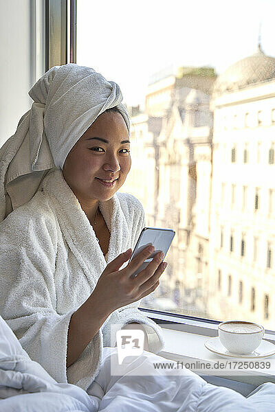 Junge Frau im Bademantel hält Handy am Fenster eines Hotels