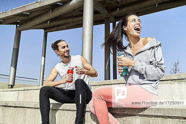 Lächelnder Mann mit Wasserflasche  der neben einer lachenden Freundin auf den Stufen eines sonnigen Tages sitzt