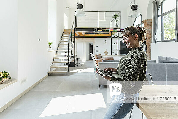Lächelnde Geschäftsfrau  die einen Laptop benutzt und sich zu Hause auf einen Tisch stützt