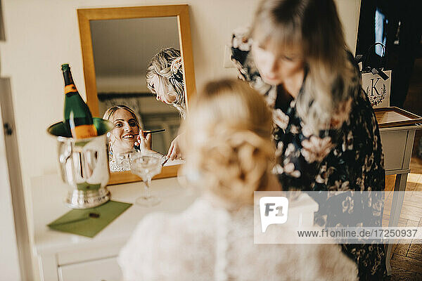 Bride looking in mirror while beautician applying make-up during wedding