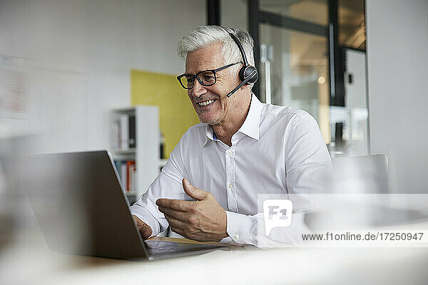 Lächelnder Geschäftsmann mit Headset gestikuliert bei einem Videogespräch auf einem Laptop im Büro