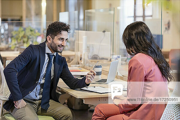 Lächelnder männlicher Fachmann  der einer Unternehmerin einen Stift anbietet  während sie am Schreibtisch in einem Coworking-Büro sitzt