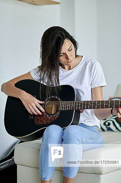 Mittlere erwachsene Frau spielt Gitarre  während sie zu Hause auf dem Sofa sitzt