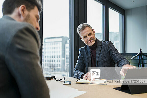 Lächelnde männliche Fachkraft  die mit einem Kollegen am Arbeitsplatz über ein digitales Tablet diskutiert