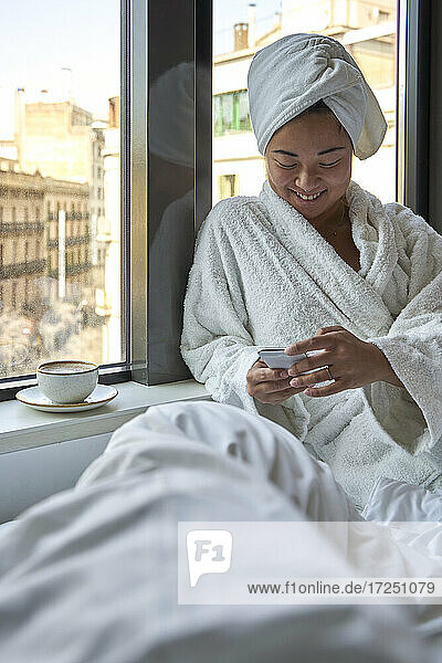 Lächelnde Frau  die ein Smartphone benutzt  während sie auf einem Hotelbett sitzt