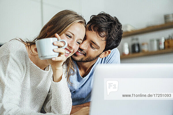 Happy man embracing girlfriend looking at laptop in kitchen