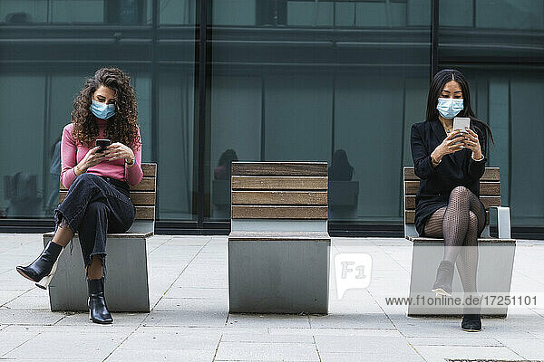 Junge Frauen mit Gesichtsschutz  die ein Mobiltelefon benutzen  während sie mit sozialem Abstand auf einer Bank sitzen