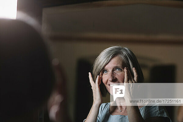 Lächelnde Frau mit Kopf in den Händen  die zu Hause in den Spiegel schaut