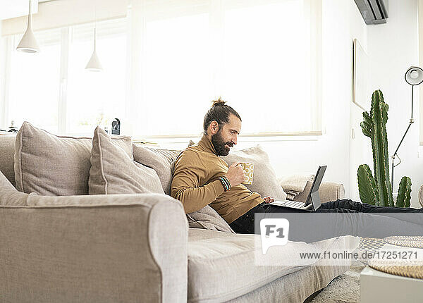 Mann mit Kaffeetasse und Tablet-PC  während er zu Hause auf dem Sofa sitzt