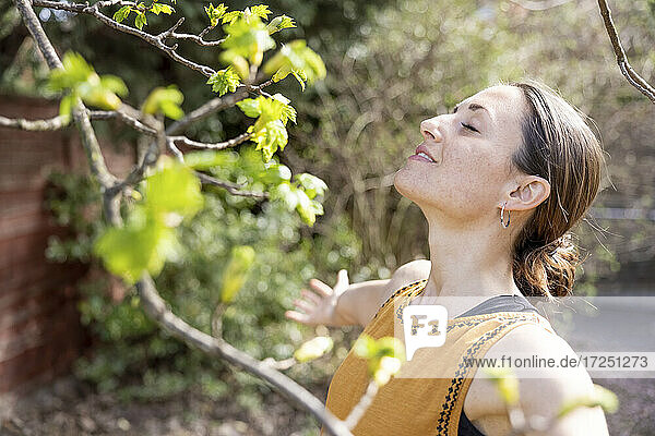 Entspannte Frau mit geschlossenen Augen an einem Zweig im Garten stehend
