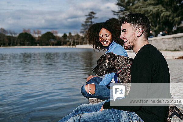 Lächelndes junges Paar sitzt mit Hund am See an einem sonnigen Tag