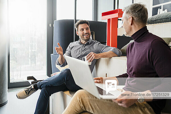 Lächelnder Geschäftsmann im Gespräch mit einem männlichen Kollegen mit Laptop am Arbeitsplatz
