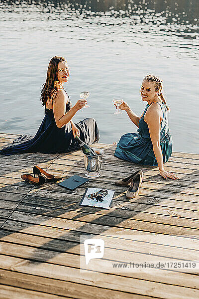 Schöne lächelnde weibliche Event-Planer sitzen mit Champagner auf Steg über See