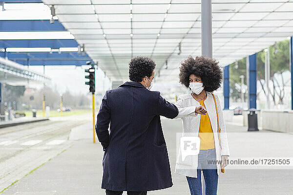 Frau mit Schutzmaske stößt männlichen Freund am Bahnhof mit dem Ellbogen an